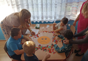 Dzieci z grupy 1 rozdmuchują przez słomki bibułę z emotki przedstawiającej smutek.