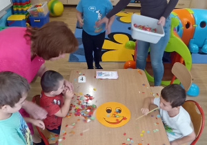 Dzieci z grupy 4 rozdmuchują przez słomki bibułę z emotki przedstawiającej radość.